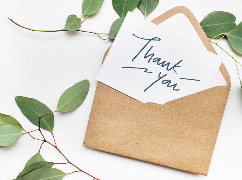 White thank you card in a brown envelope surrounded by green leaves, sending you a gratitude message this holiday season.