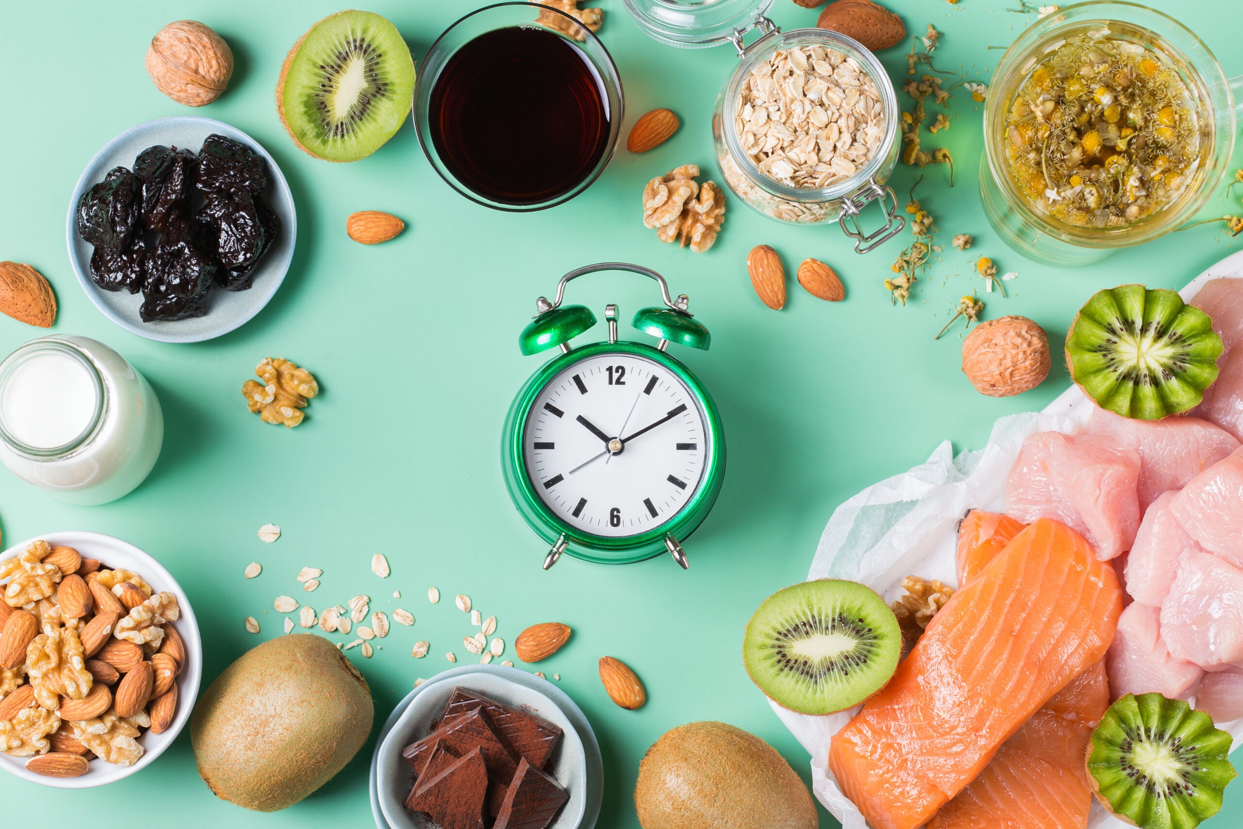 Flat lay of a green clock surrounded by salmon, kiwi, nuts, oatmeal, and tart cherry juice. Showing that intermittent fasting and nutrition can increase mitochondrial health and energy production.