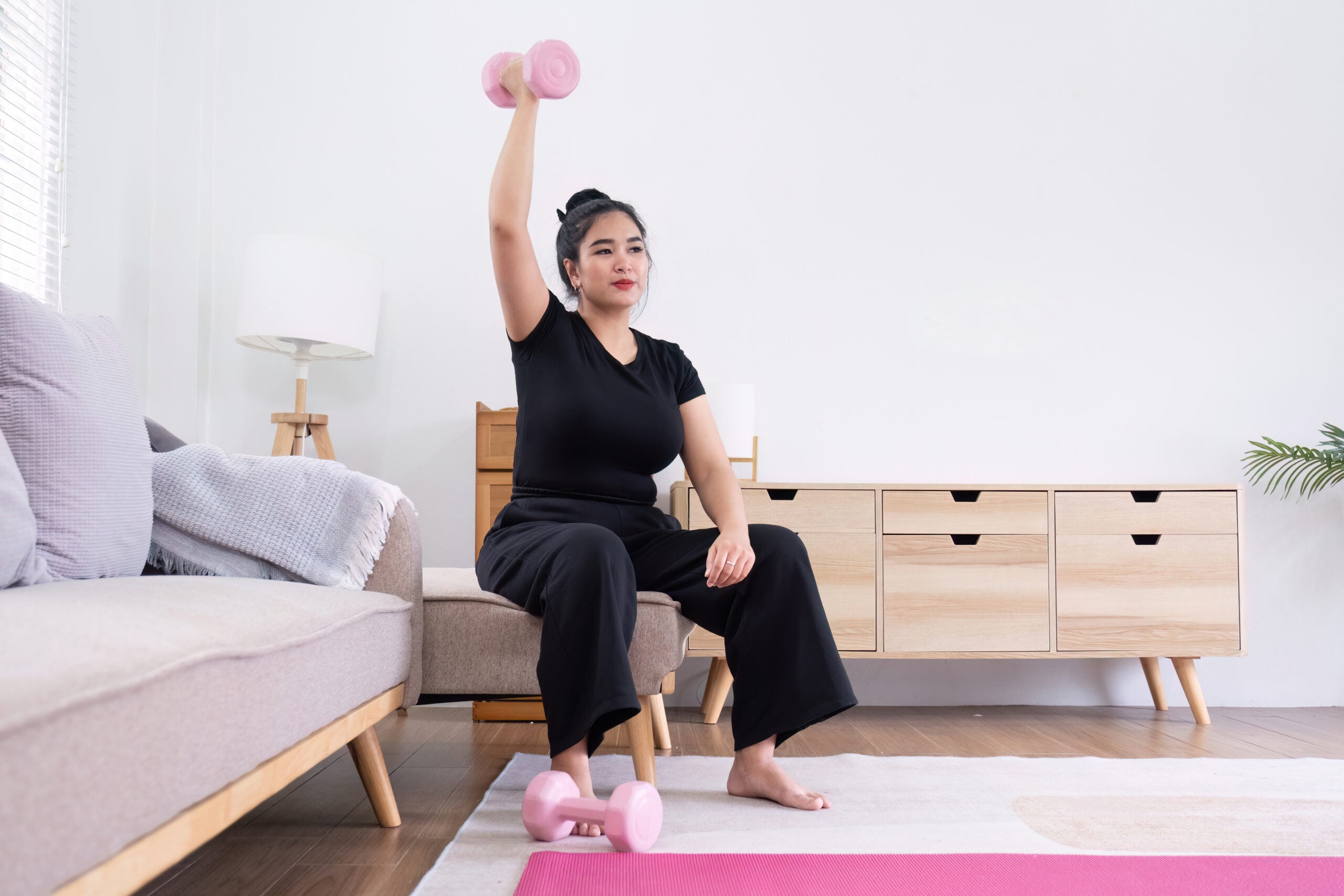 Young Asian working out with pink dumbbells in her living room and using weight lifting to help reduce her toxin exposure and increase her bone health.