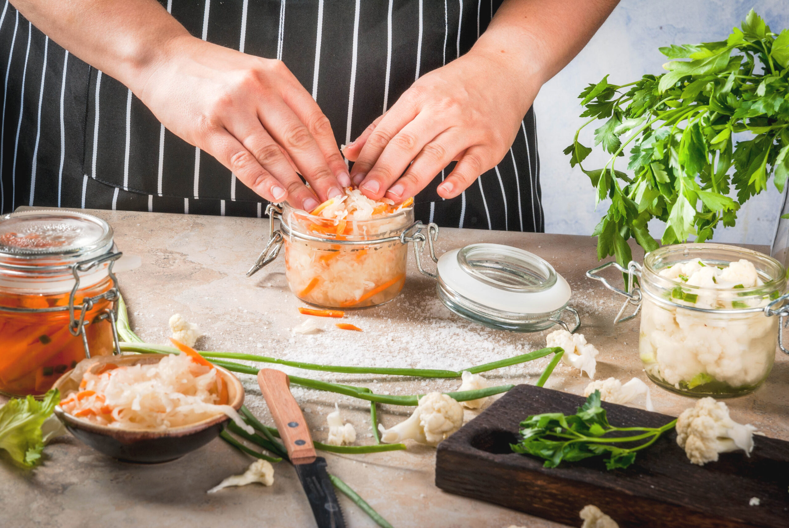 Close up of a woman's hands putting kimchi in a glass surrounded by herbs and gut health foods for balancing the gut-brain axis.